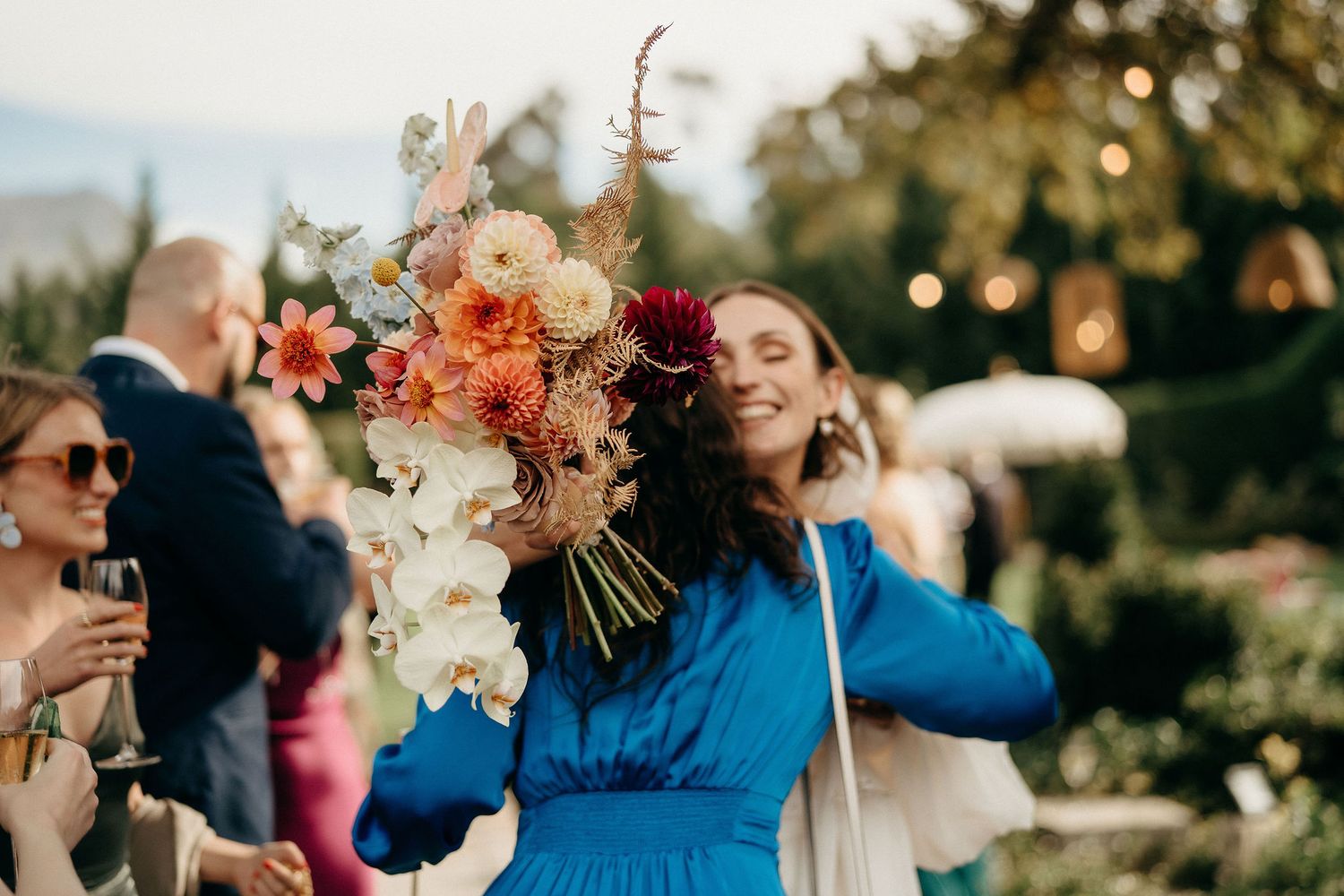Person in bright blue dress celebrates outdoors holding a vibrant floral bouquet with orange and white flowers.