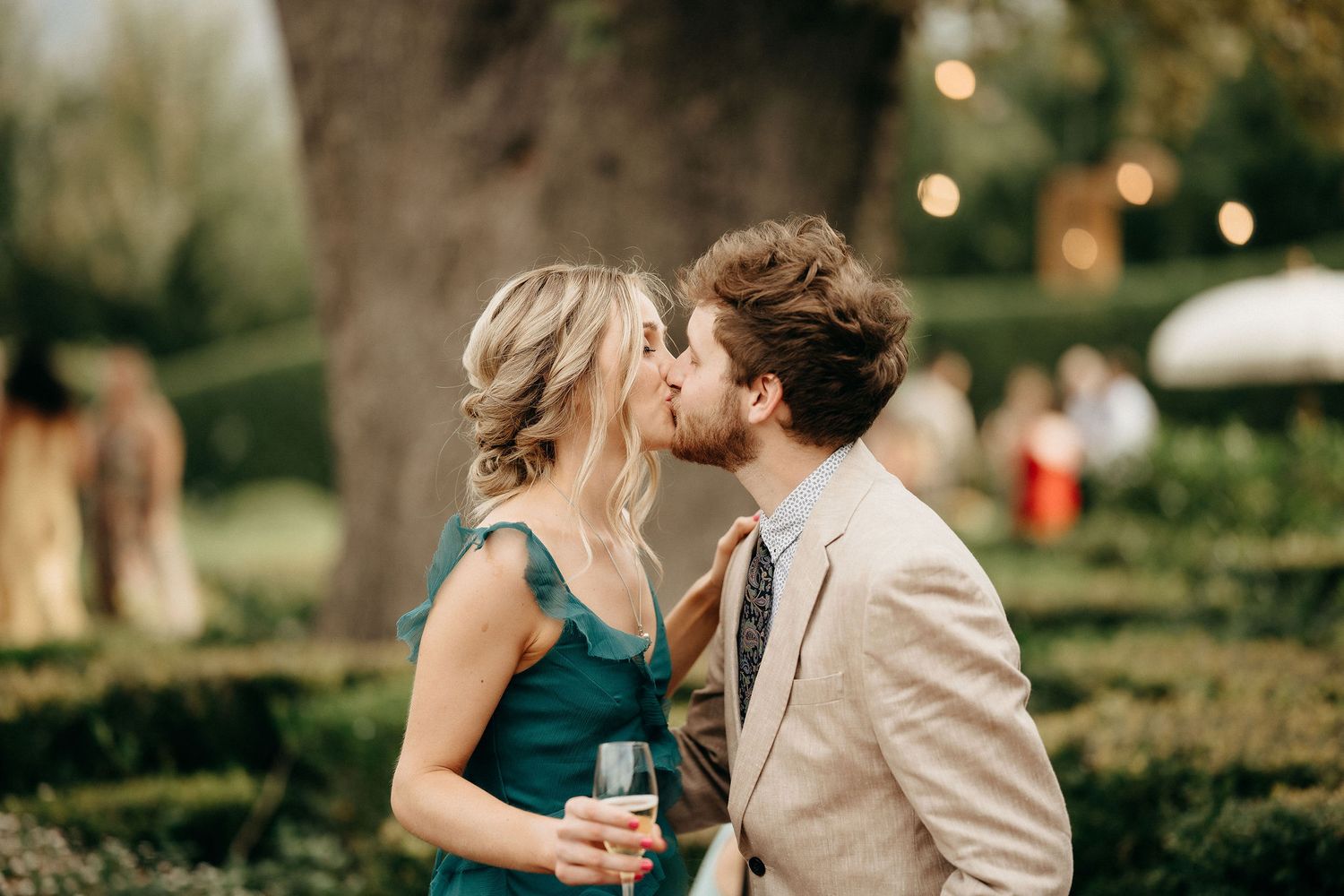A couple shares a romantic kiss in a garden setting with soft bokeh lights in the background.