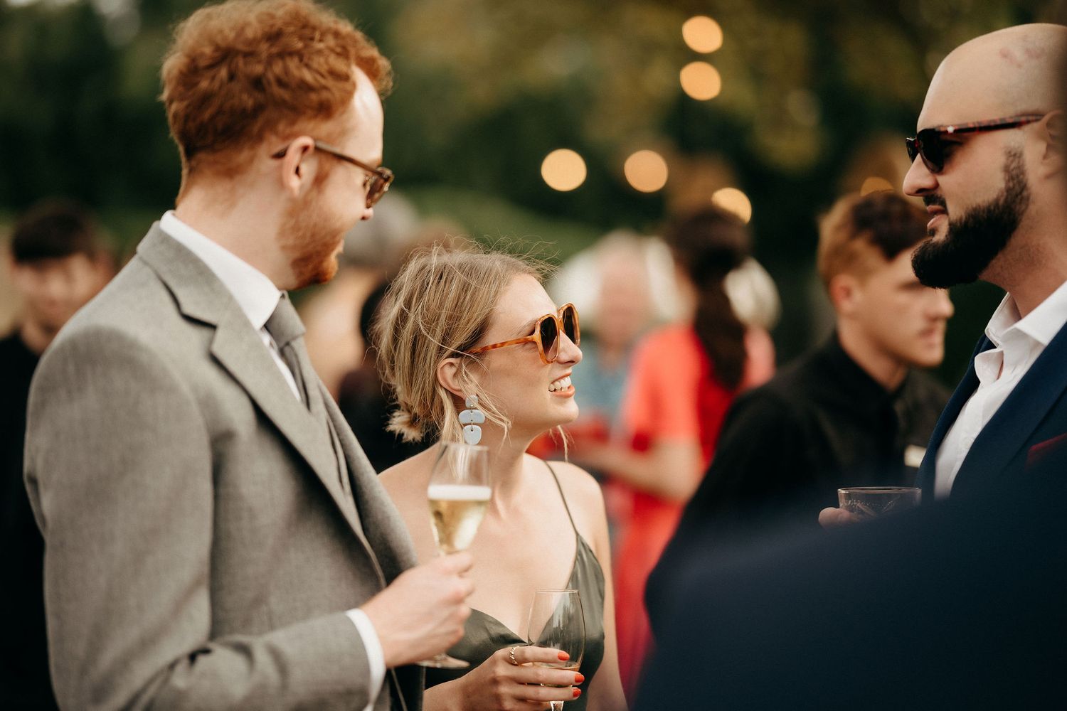 Guests in stylish attire enjoying drinks and conversation at an outdoor evening event with bokeh lighting in background.