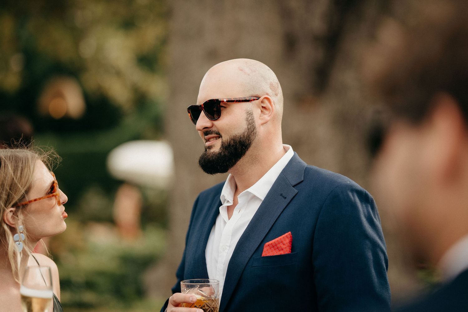 Stylish guest in navy suit with red pocket square and sunglasses smiles while holding drink at outdoor event.