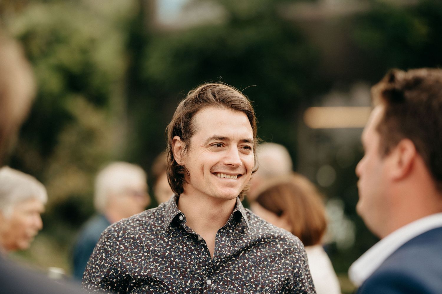 A young person in a patterned button-down shirt smiles warmly at an outdoor social gathering.