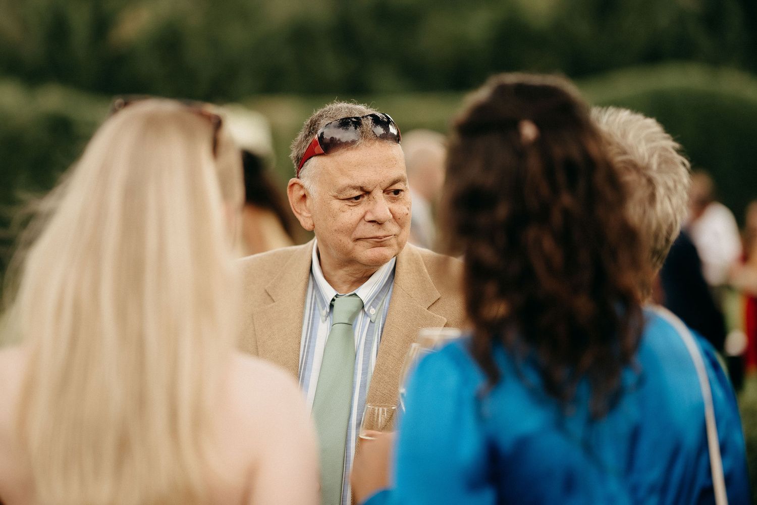 A person in a beige suit and light blue tie stands among others during an outdoor gathering.