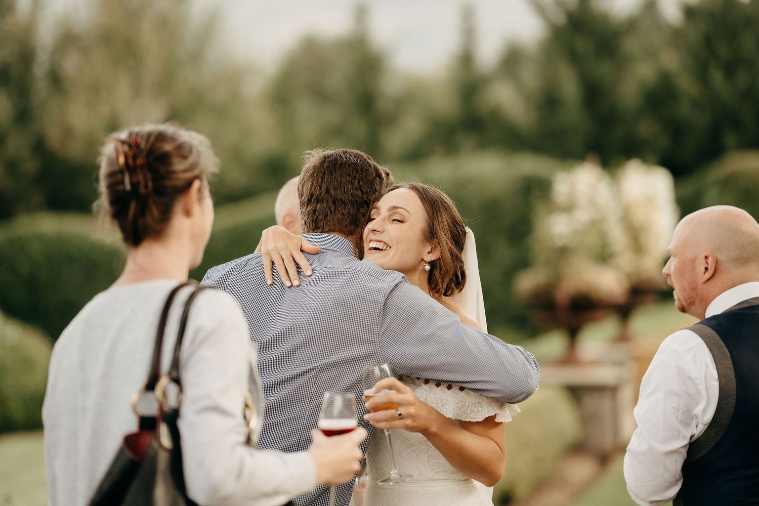 People embrace and celebrate at an outdoor wedding reception in a garden setting with wine glasses in hand.
