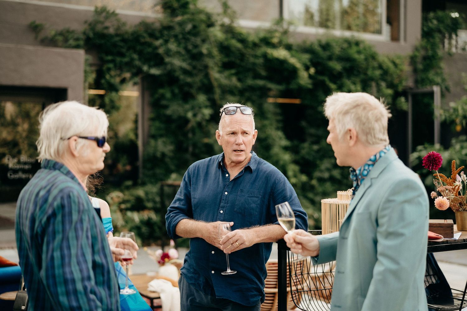 Three people engaged in casual conversation on an outdoor patio with greenery and wooden chairs in the background.