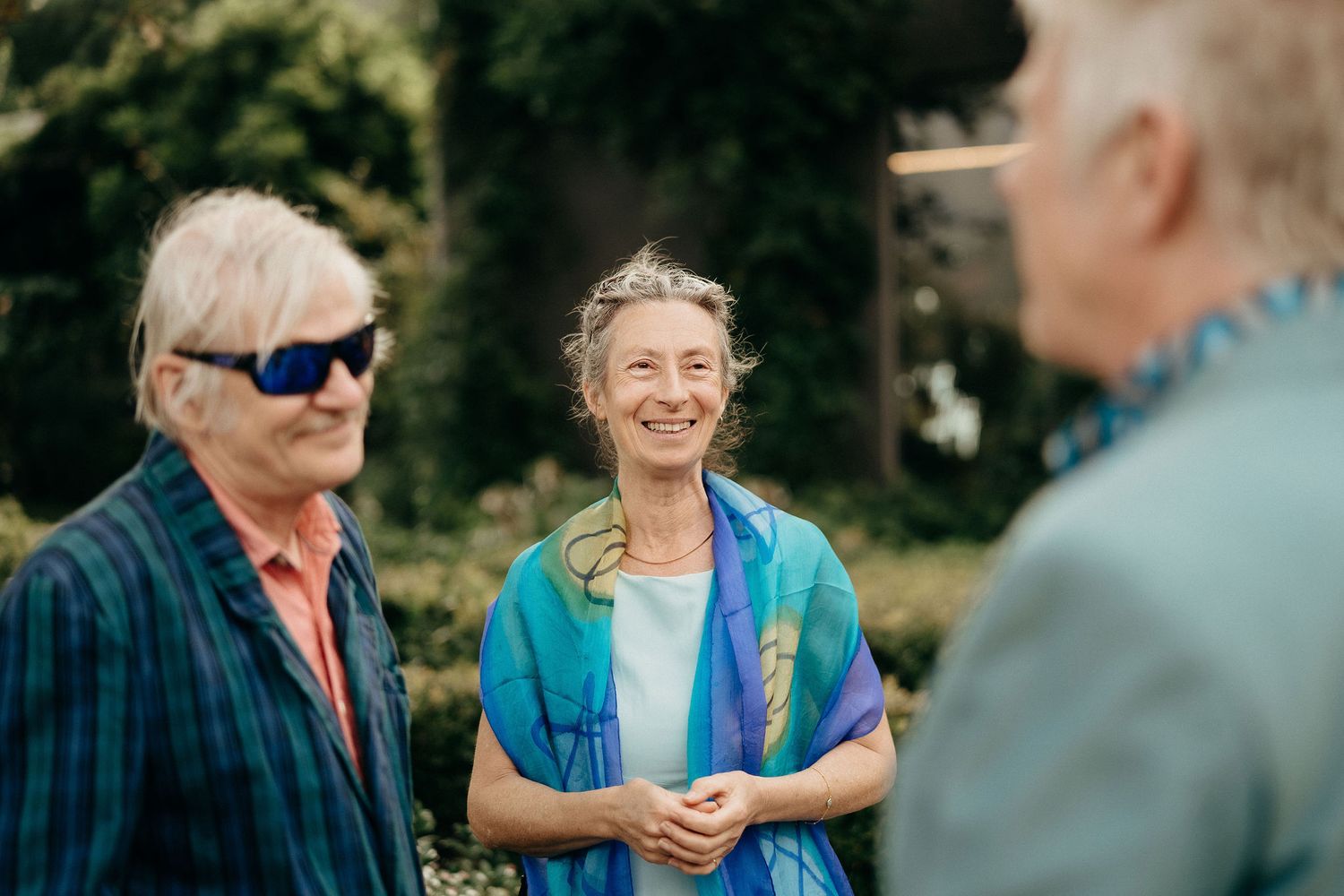 Three people engaged in friendly conversation outdoors in a garden setting on a sunny day.