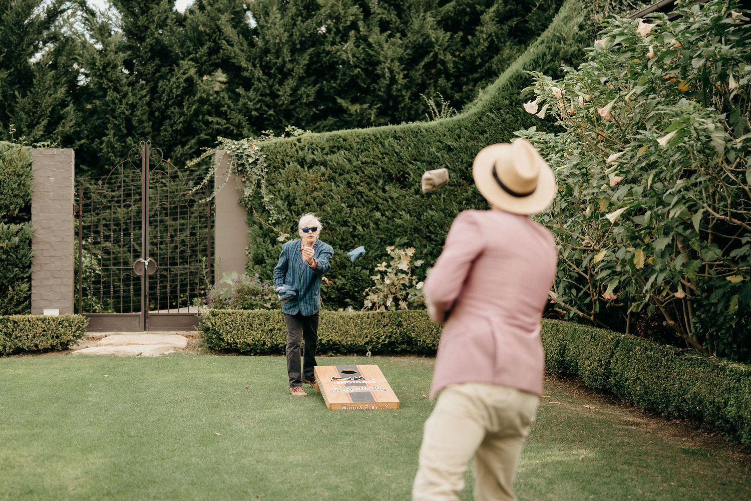 Two people playing cornhole on a manicured lawn with hedges and stone pillars in a garden setting.