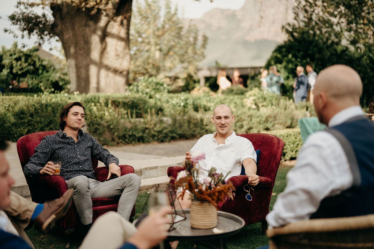 A group gathers for conversation around outdoor patio furniture in a lush garden setting at dusk.
