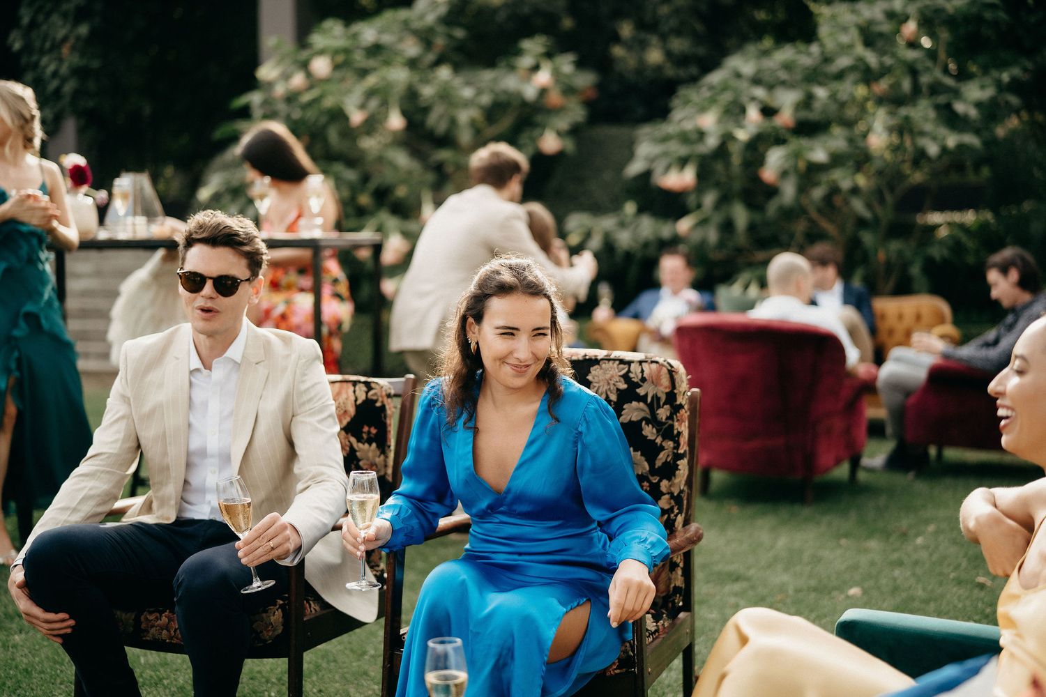 Guests in formal attire enjoy an outdoor garden party with scattered seating arrangements on a sunny day.