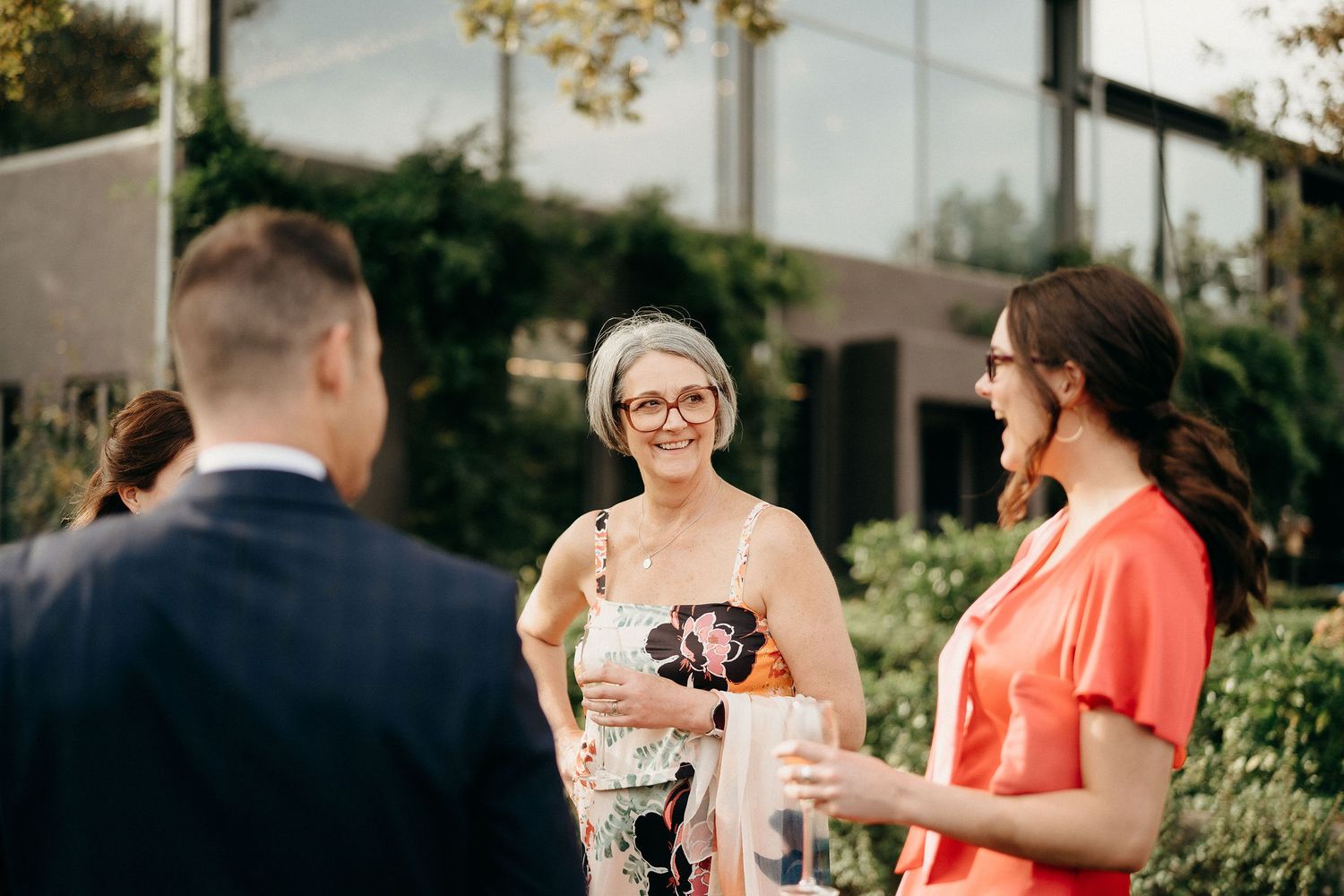Three people engaged in conversation at an outdoor event with greenery and buildings in the background.