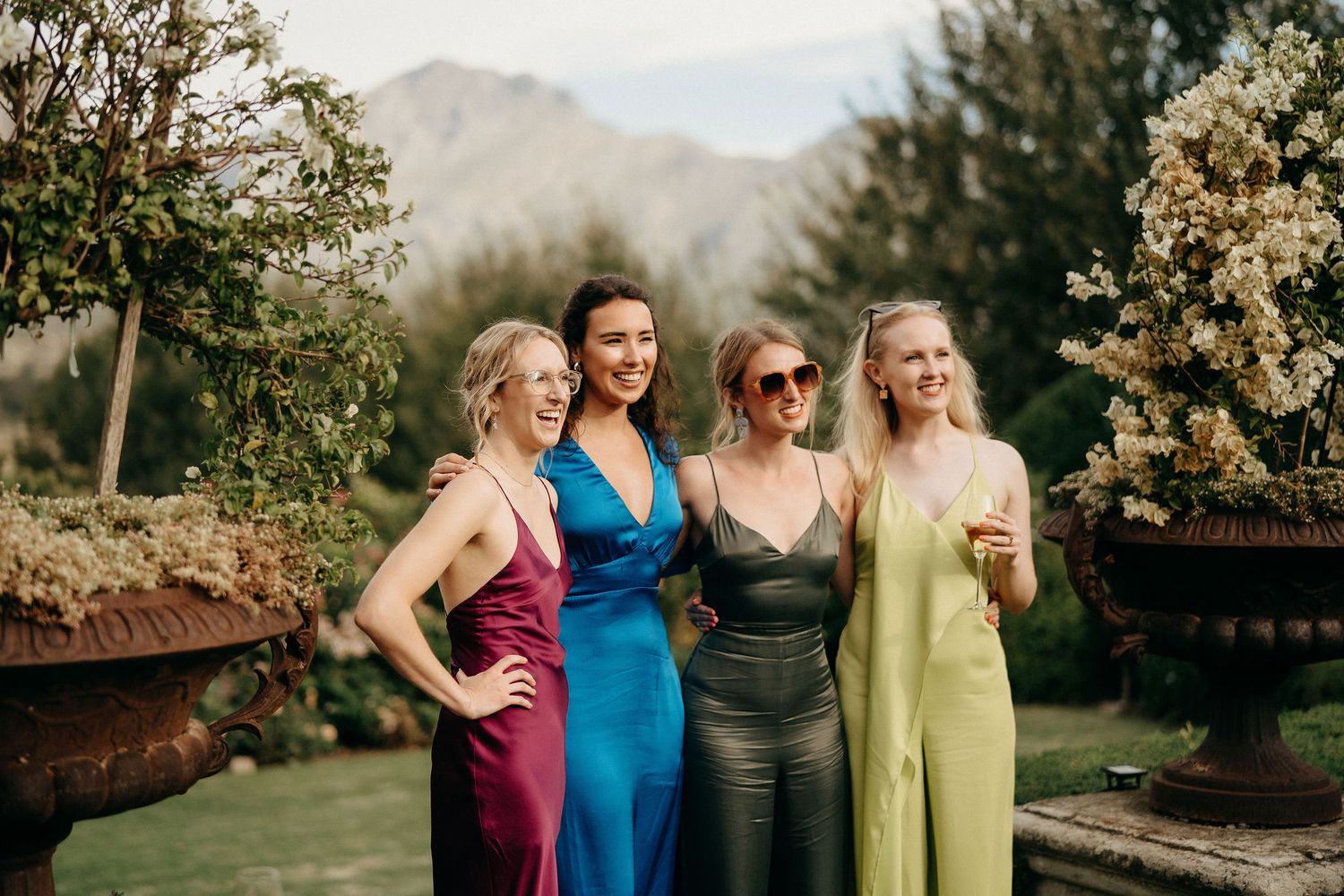Four people pose together at an outdoor garden venue with mountains in the background wearing elegant evening dresses.