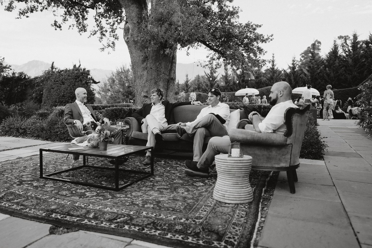 Black and white photograph showing people relaxing on outdoor patio furniture under a large tree at a social gathering.