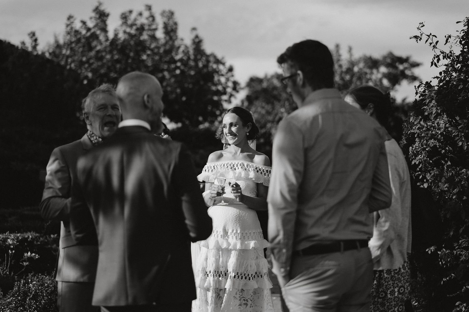 A black and white photograph of an outdoor wedding ceremony with the bride and officiant surrounded by trees and foliage.
