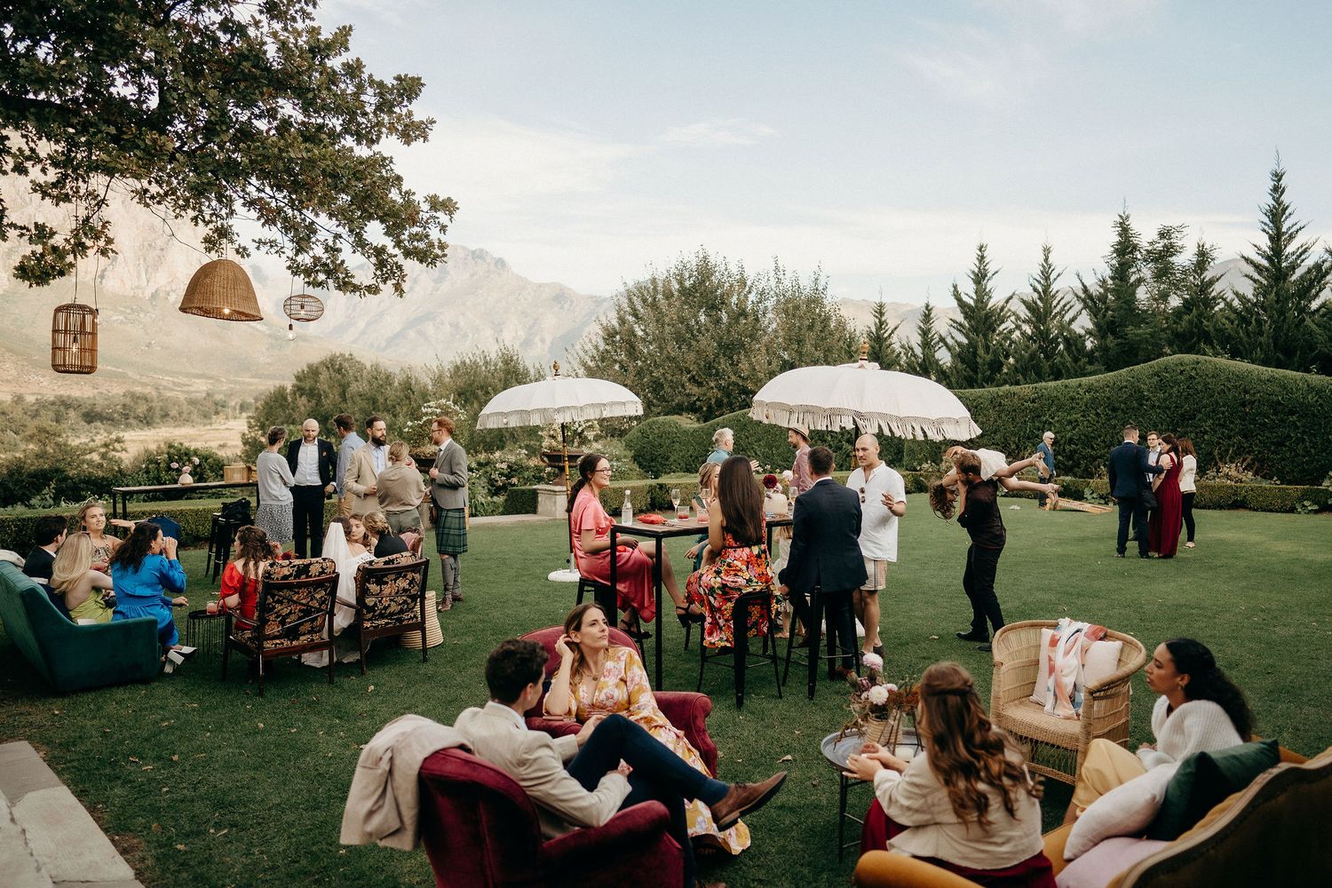 Outdoor garden party with white umbrellas, guests lounging on chairs and blankets during a sunny afternoon gathering.