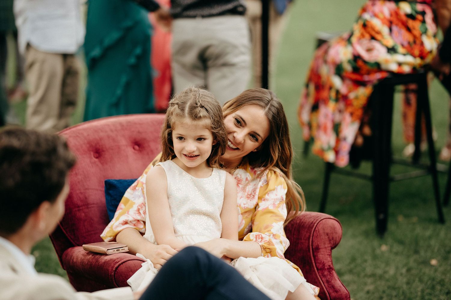 Two people share a tender moment while sitting together on a pink vintage armchair outdoors on grass.