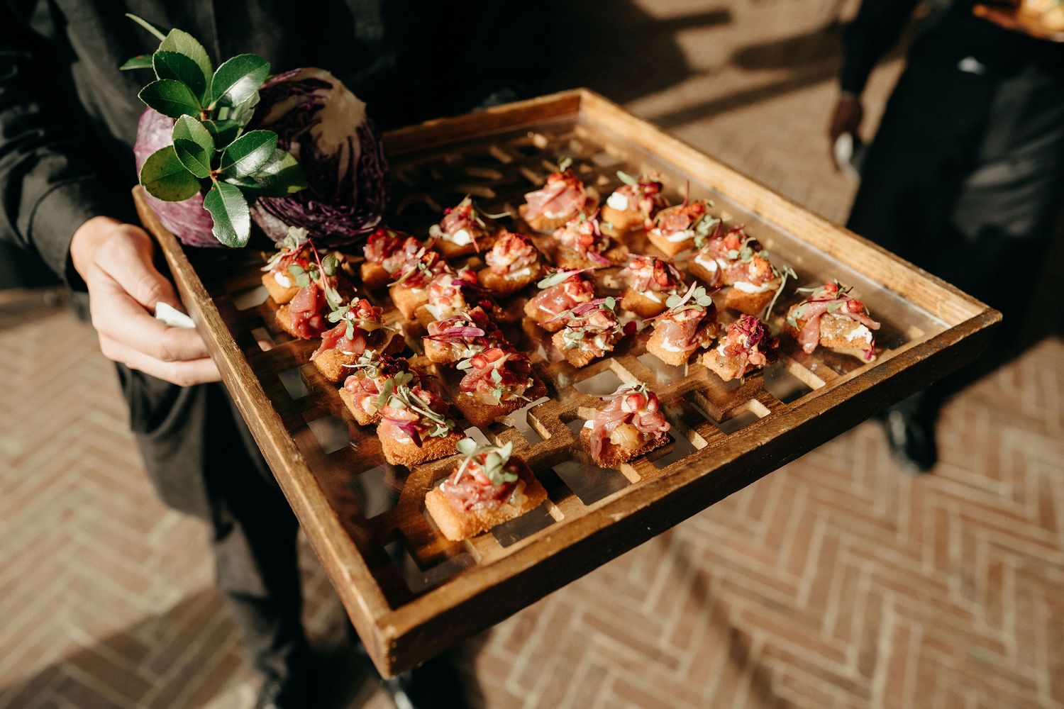 A wooden serving tray filled with appetizers garnished with fresh herbs being presented at a catered event.