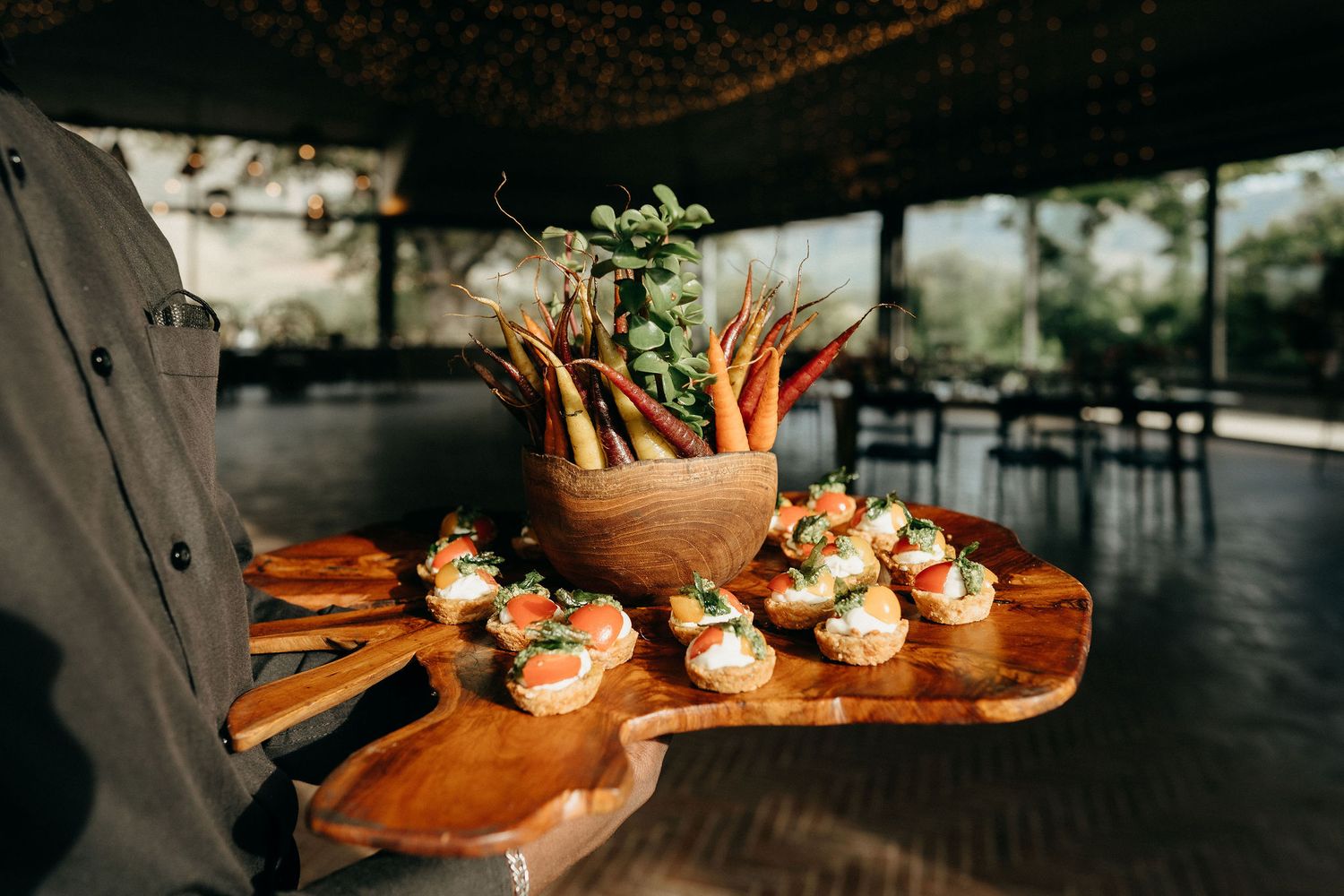 A wooden serving board with baby carrots and appetizers being presented at a restaurant's covered outdoor dining area.