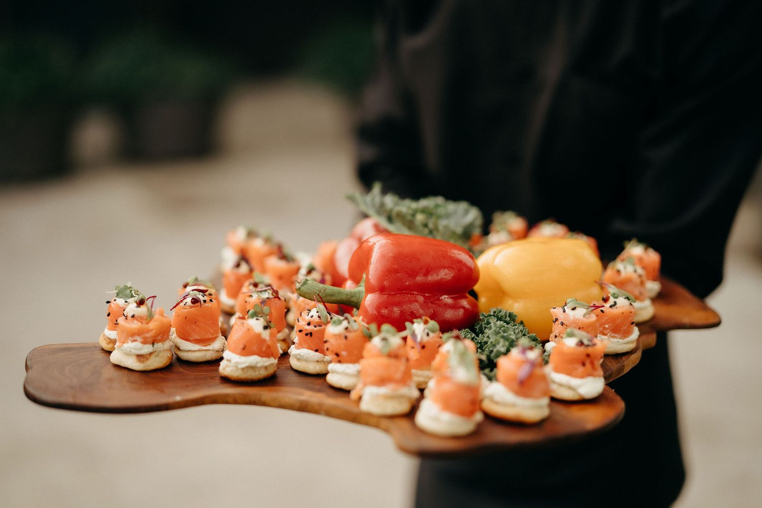 An elegant wooden serving platter displays assorted gourmet appetizers with salmon and garnishes at a catered event.
