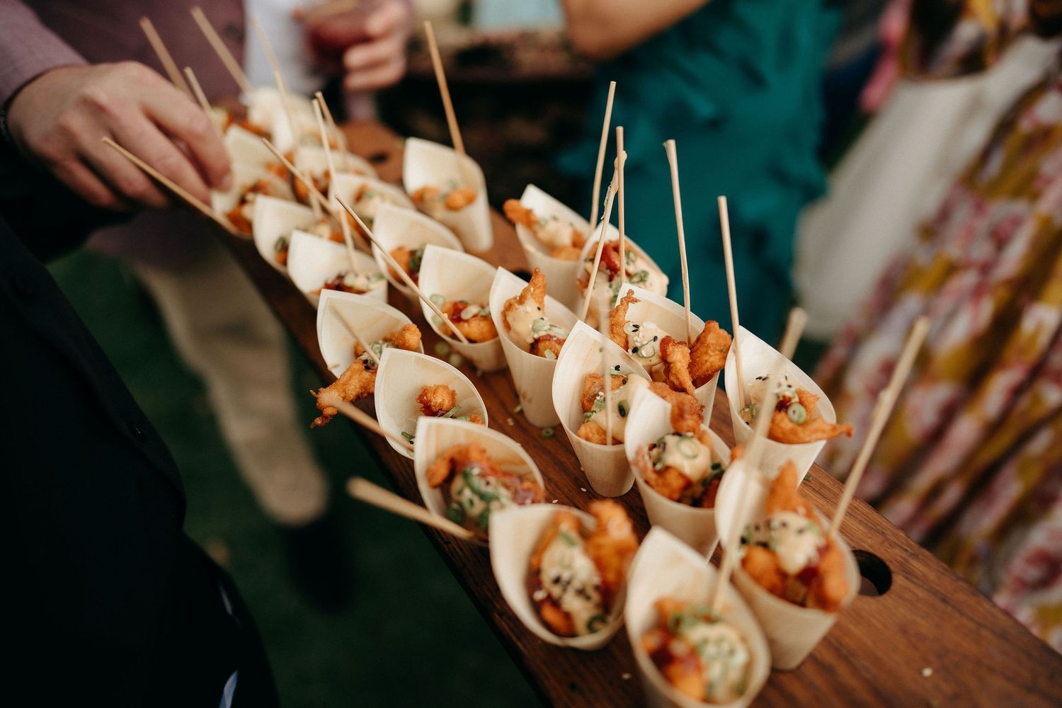 Small paper cones filled with appetizers arranged on a wooden serving board with toothpicks at a catering event.