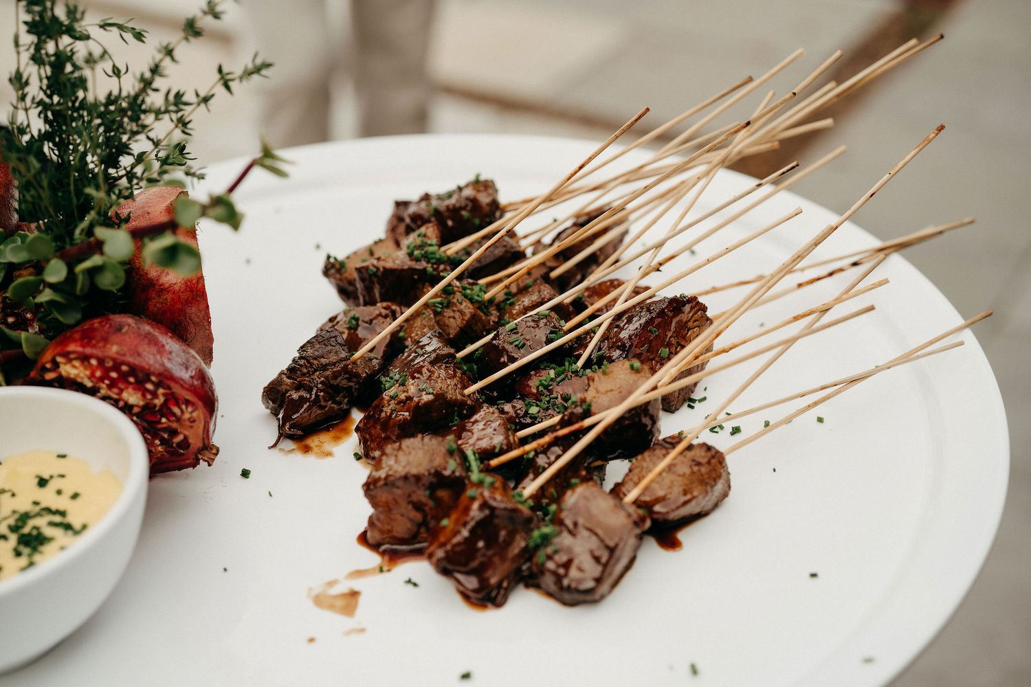 Grilled meat skewers arranged on a white plate with a side of creamy dipping sauce.