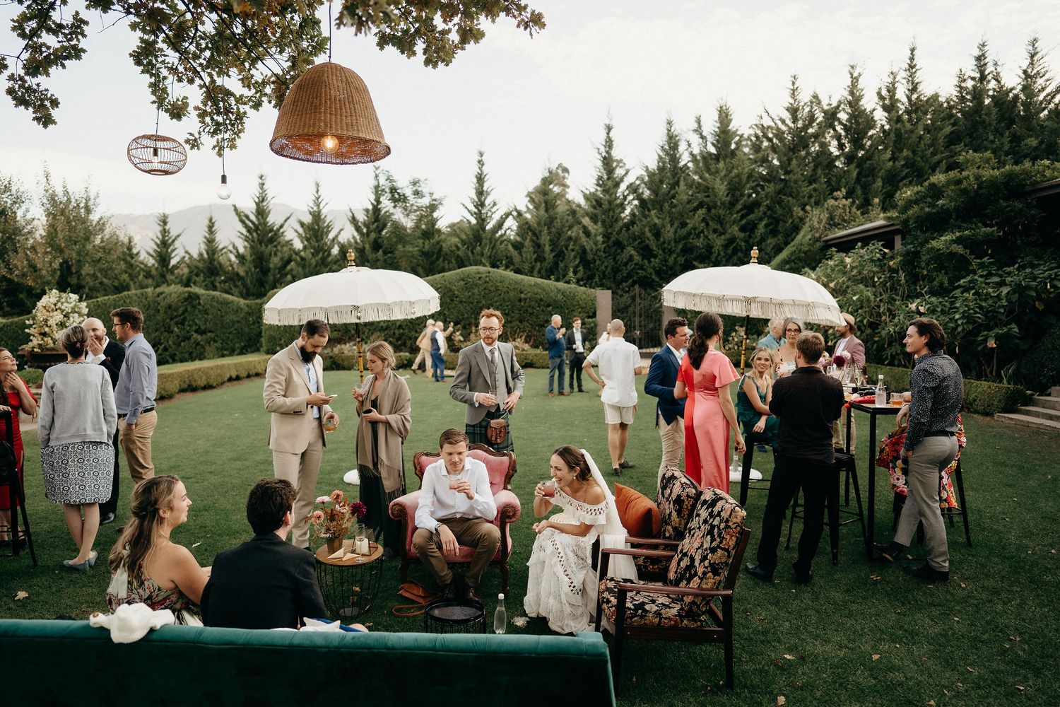 Outdoor garden party guests gather under white umbrellas and pendant lights on a green lawn bordered by evergreen trees.