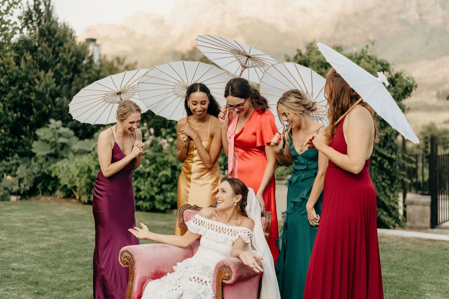 A group in colorful formal dresses pose with white parasols on a grassy lawn during an outdoor celebration.