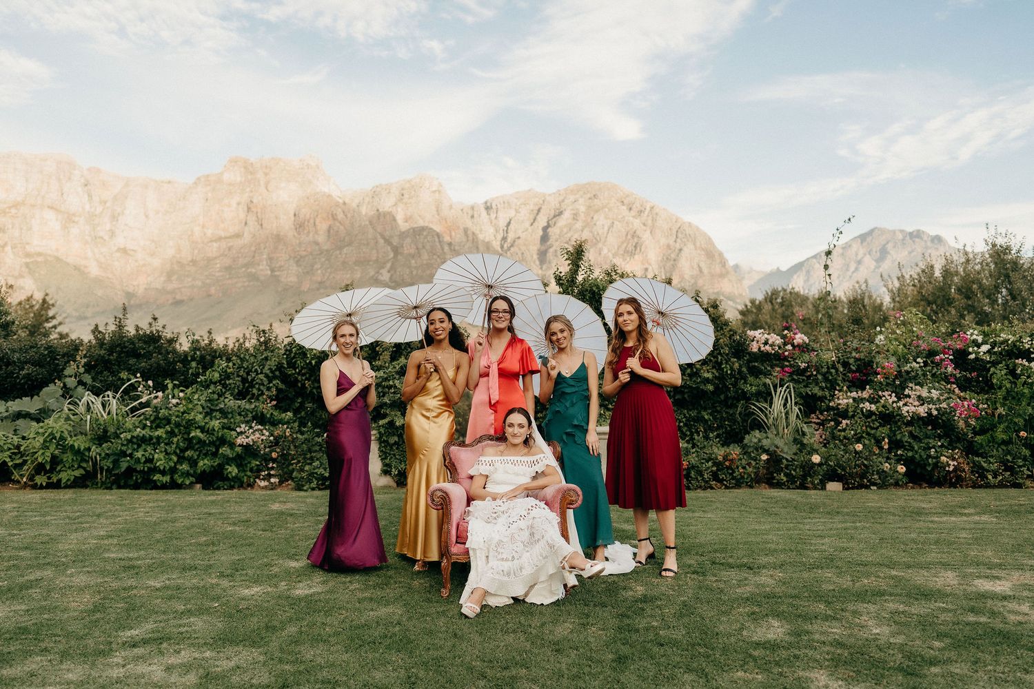 A group in formal attire poses on a lawn with parasols against a backdrop of majestic mountain peaks at golden hour.