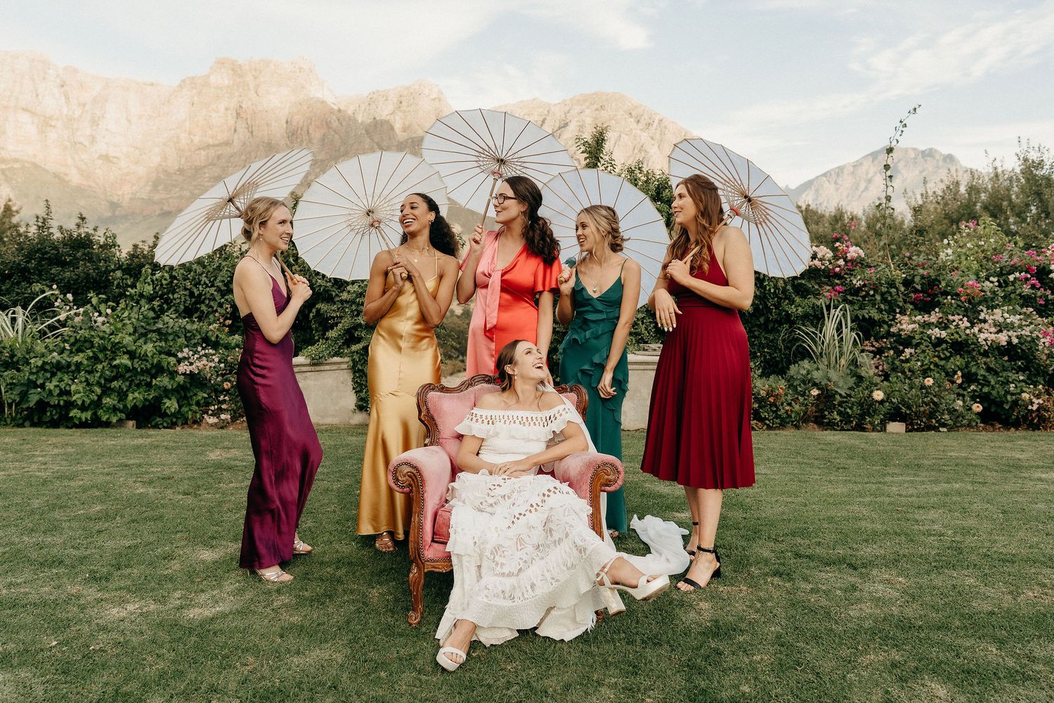 Wedding party in colorful dresses pose playfully with parasols on a grassy lawn against mountain backdrop.