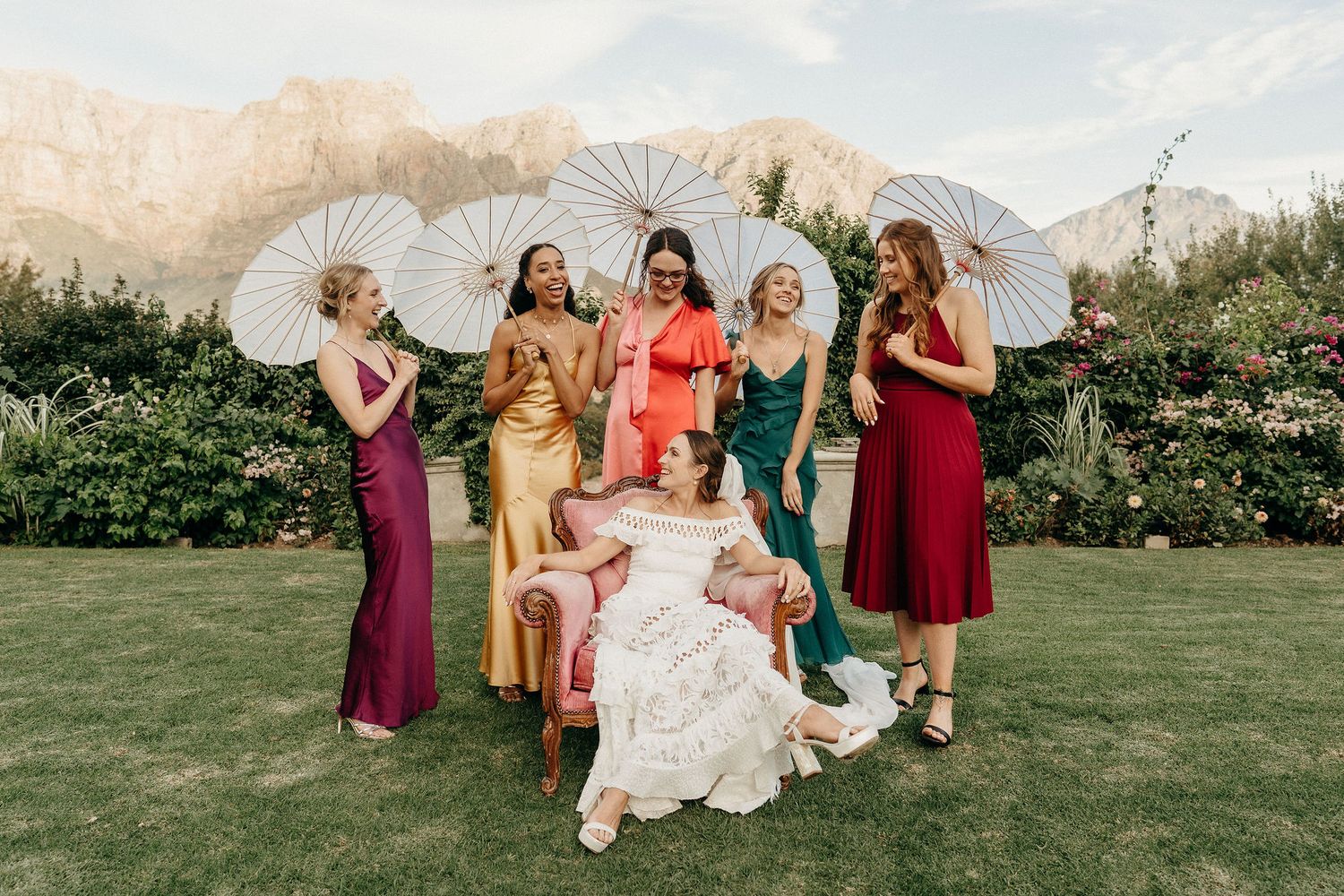 Wedding party members holding white parasols gather on a lawn wearing elegant dresses in jewel tones and white.