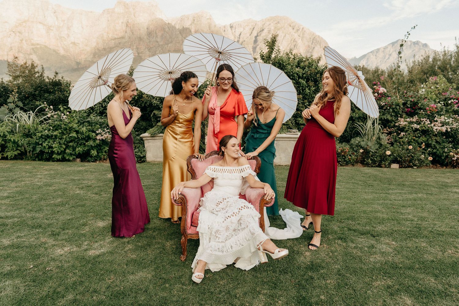 A group in colorful formal dresses pose together on a lawn with white parasols on a sunny day.
