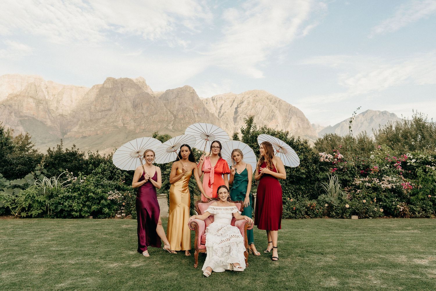 A wedding party poses with white parasols against majestic mountain backdrop on a lush green lawn.