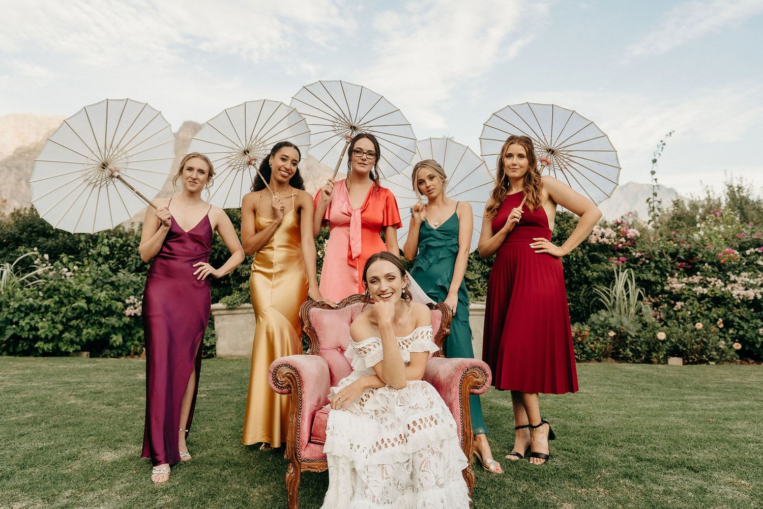 Wedding party poses with white parasols on a green lawn during an outdoor celebration.