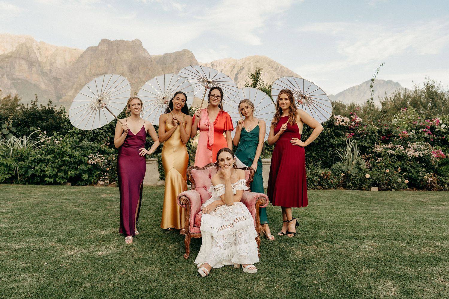 A bridal party with white parasols poses in colorful dresses against a mountainous backdrop in a garden setting.