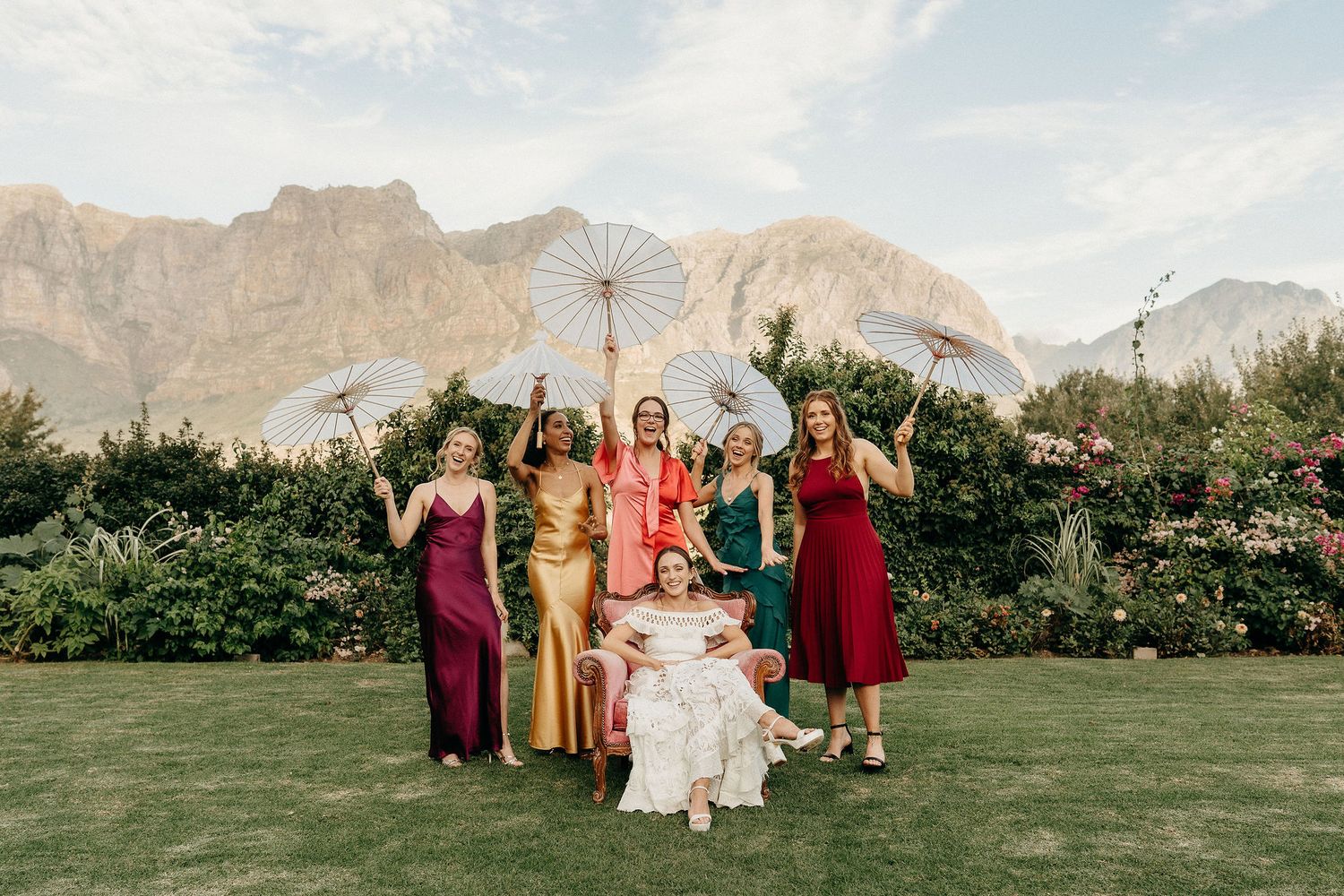 Wedding party in burgundy dresses poses on lawn with mountains and evergreen trees in background.