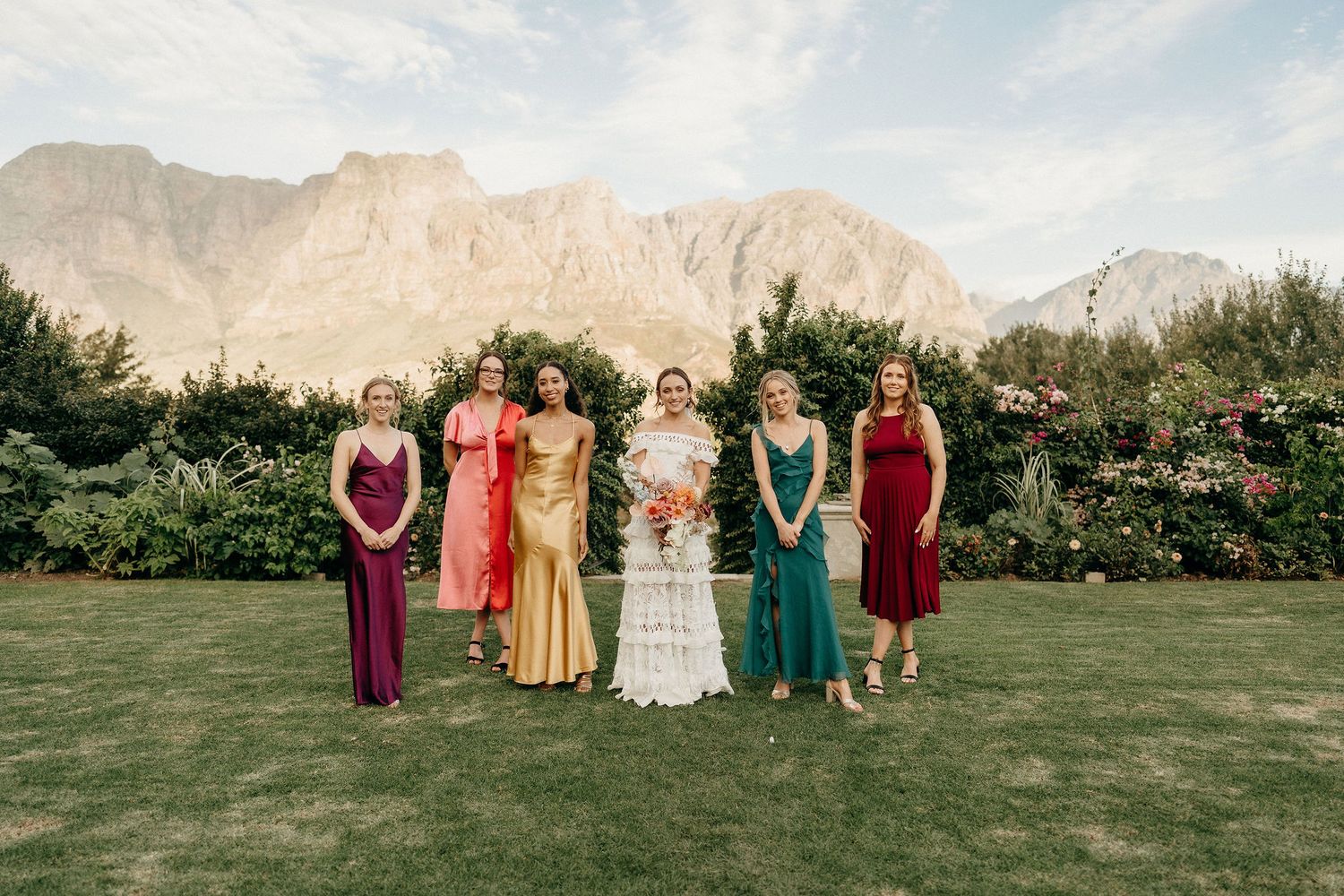 A group in colorful formal attire poses on a lawn with majestic mountain peaks rising in the background.