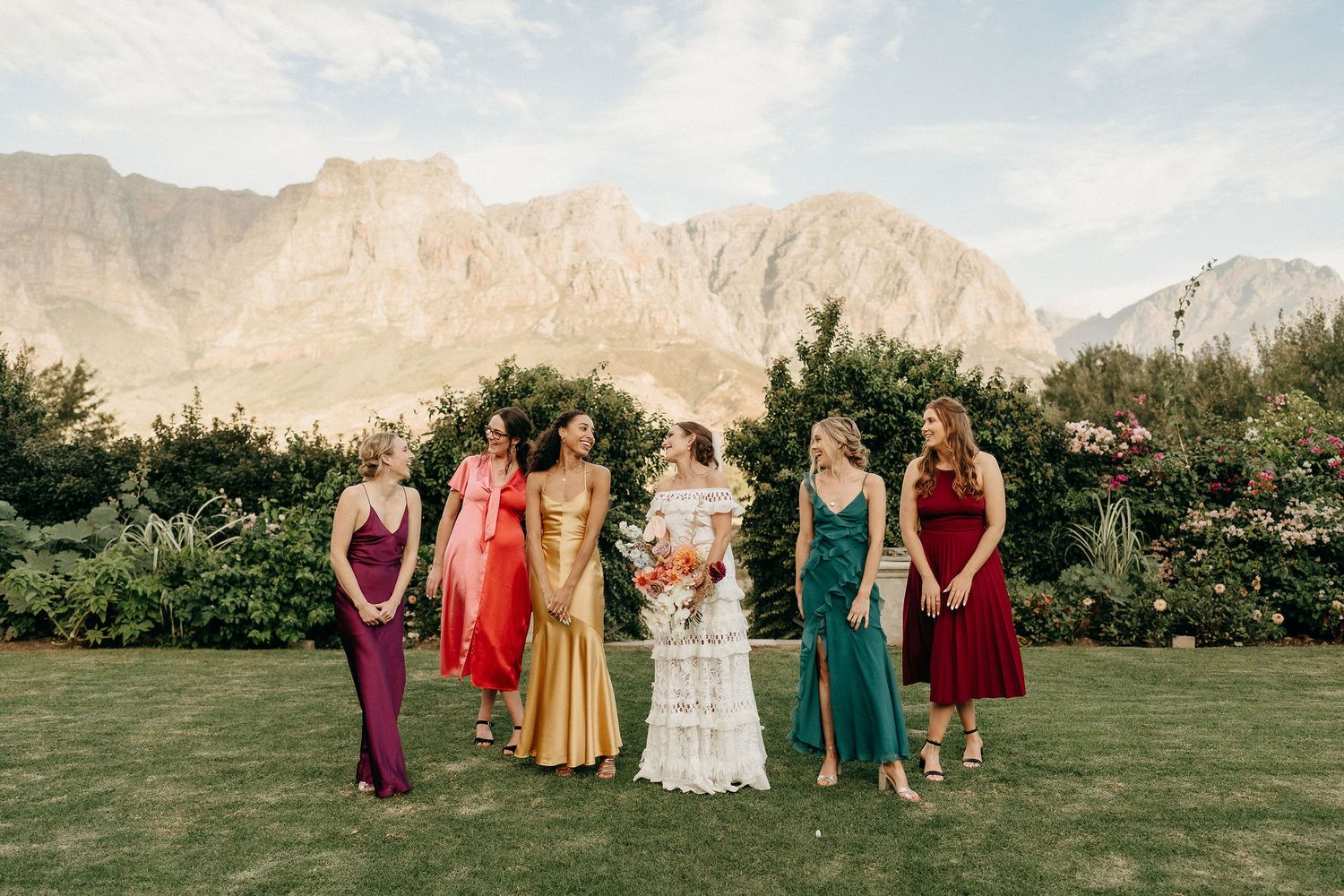 Wedding party in jewel-toned dresses poses on a lawn with dramatic mountain backdrop in Utah.