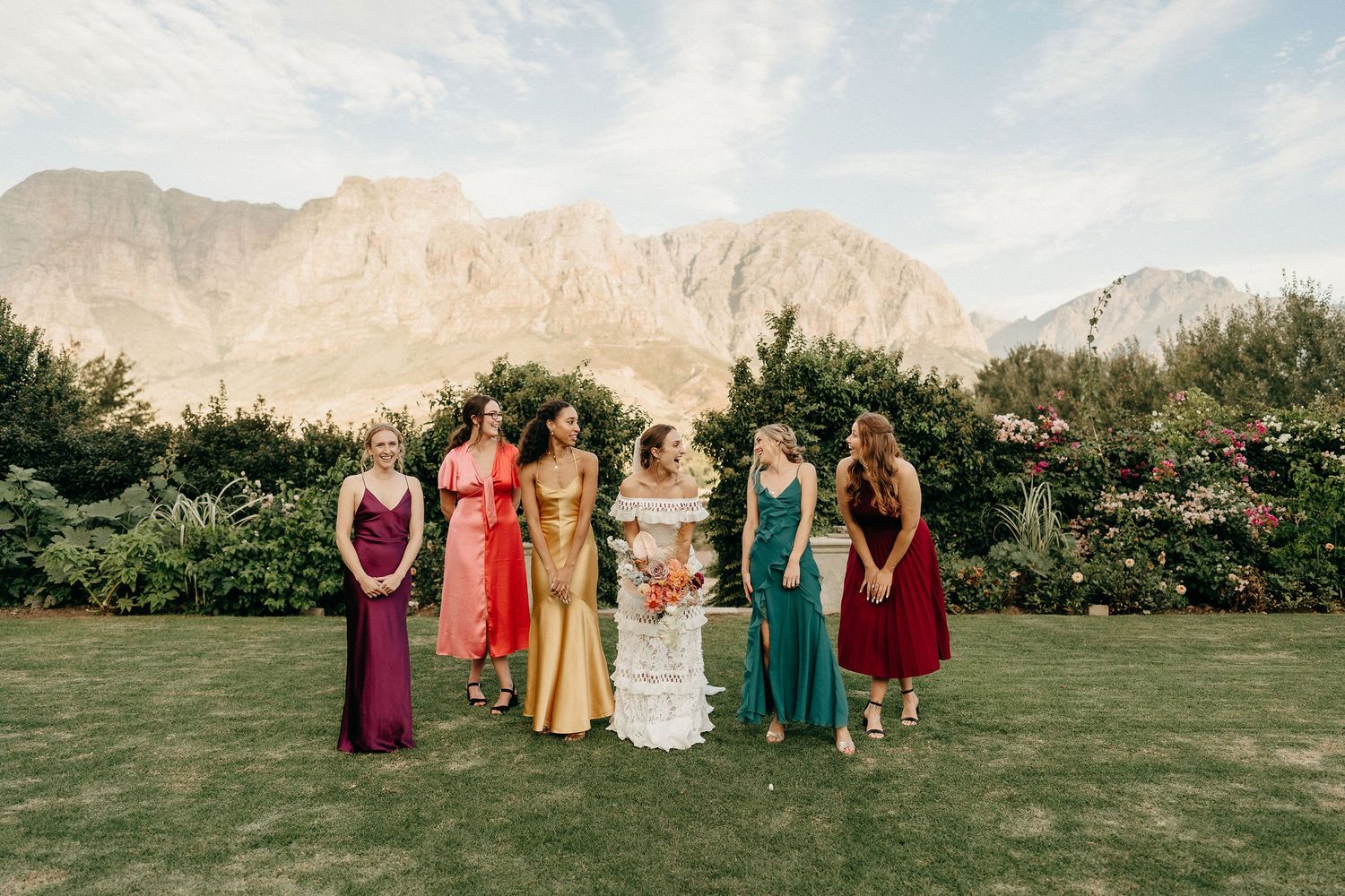 A wedding party poses on a manicured lawn against dramatic mountain peaks in a scenic outdoor setting.