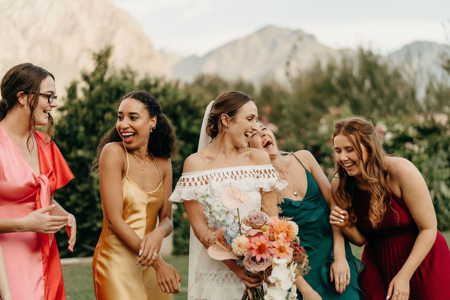Wedding party laughs together in vibrant attire with mountains and greenery in background.