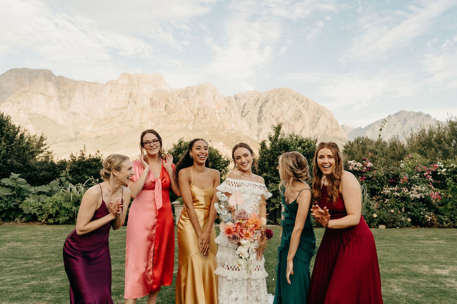 Bridesmaids in jewel-toned dresses stand together on a lawn with dramatic mountain peaks in the background.