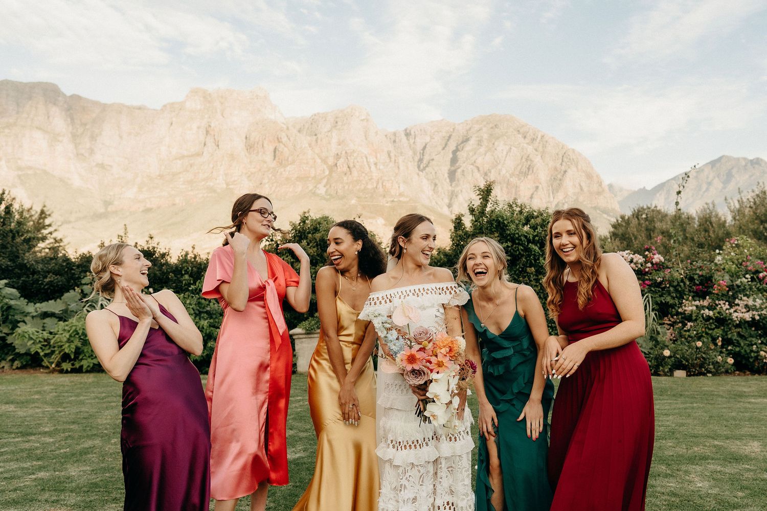 A wedding party wearing colorful dresses in pink, gold, green and burgundy laughs together with mountain backdrop.