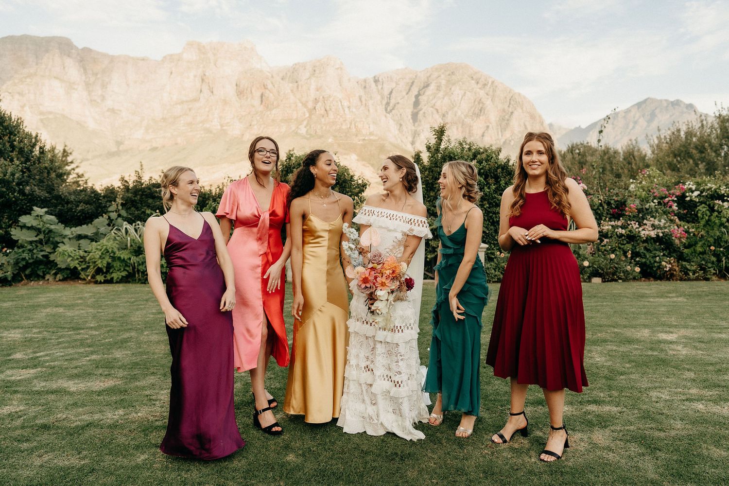 Group in colorful formal dresses including burgundy, coral, gold, white and emerald stand on grass with mountains behind them.