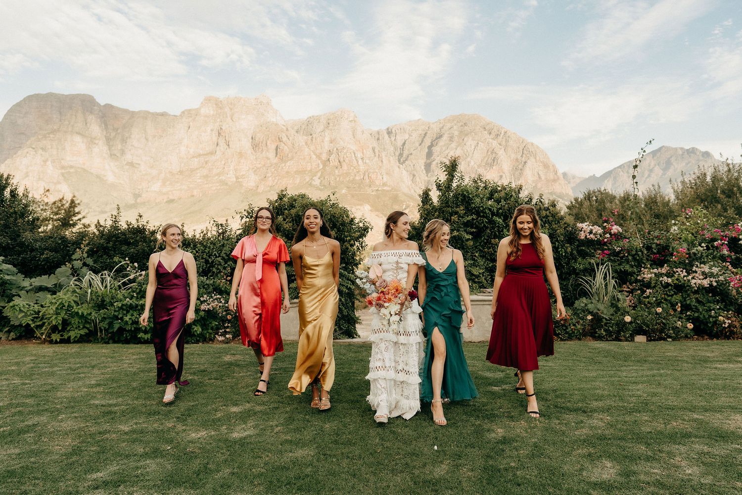 A group in colorful dresses walks together on a lawn with majestic mountains rising in the background landscape.