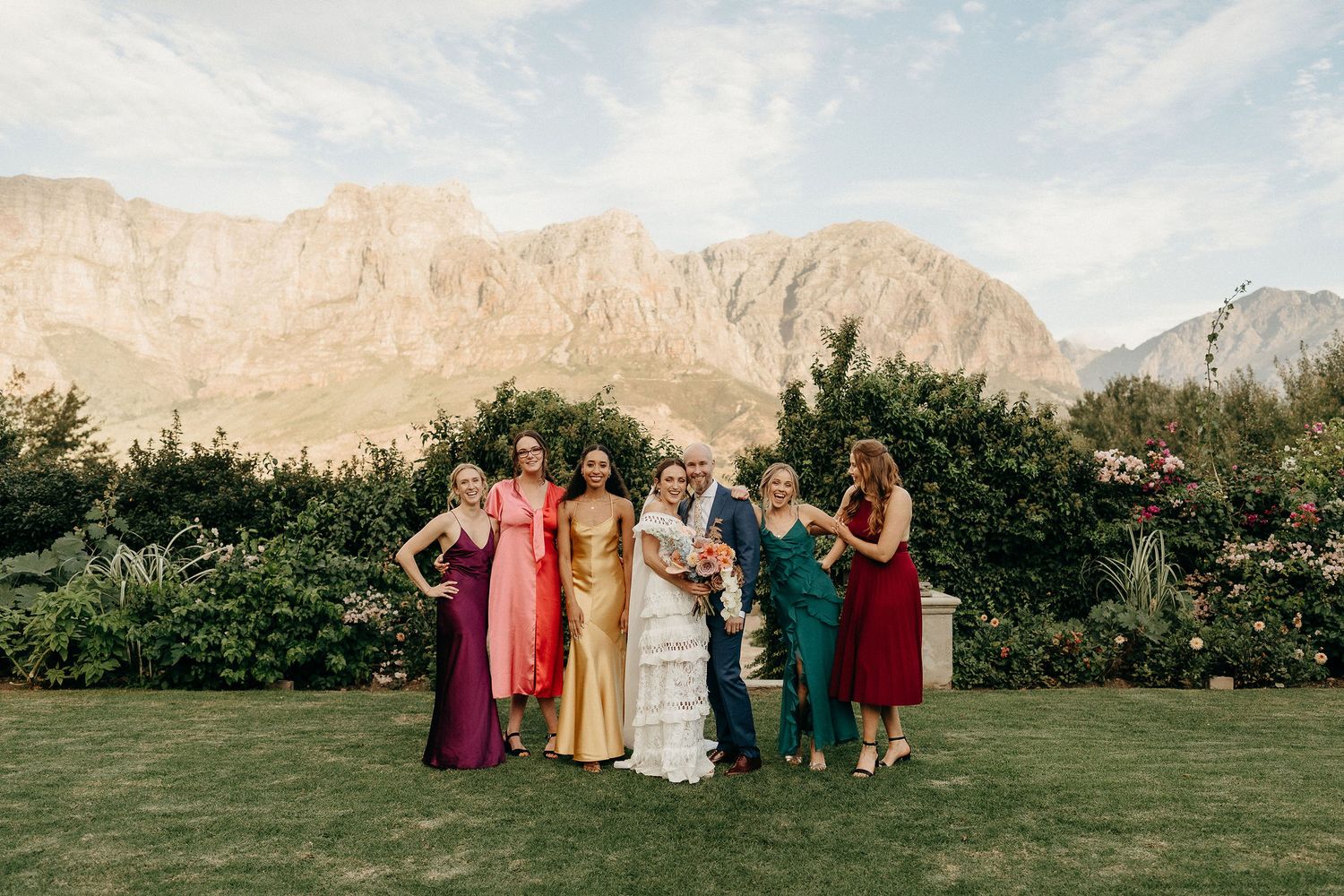 A group in formal attire poses on a lawn with dramatic mountain peaks in the background under a soft sky.