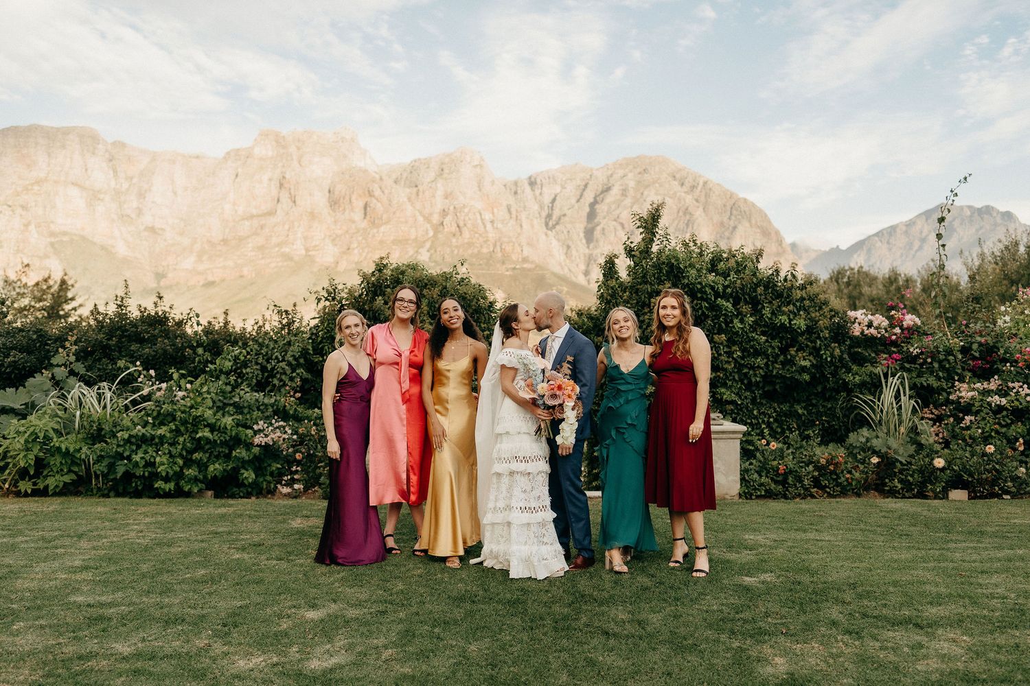 A group stands together on a green lawn with scenic mountain peaks and lush hedges in the background.
