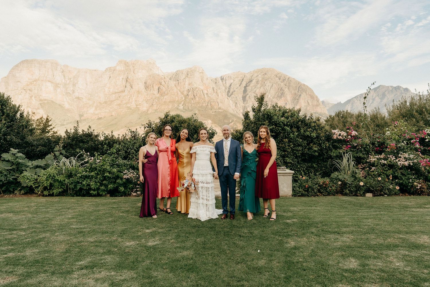 A wedding party poses on a manicured lawn with dramatic mountain peaks rising in the background.