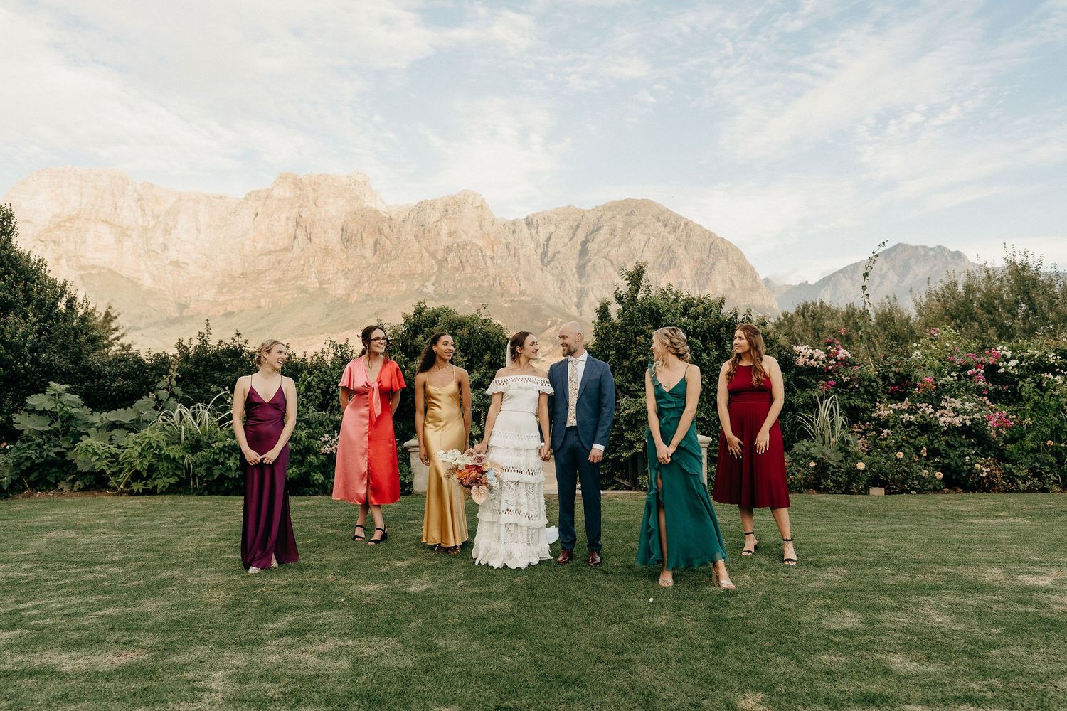 A wedding party stands on a manicured lawn with dramatic mountain peaks rising in the background.