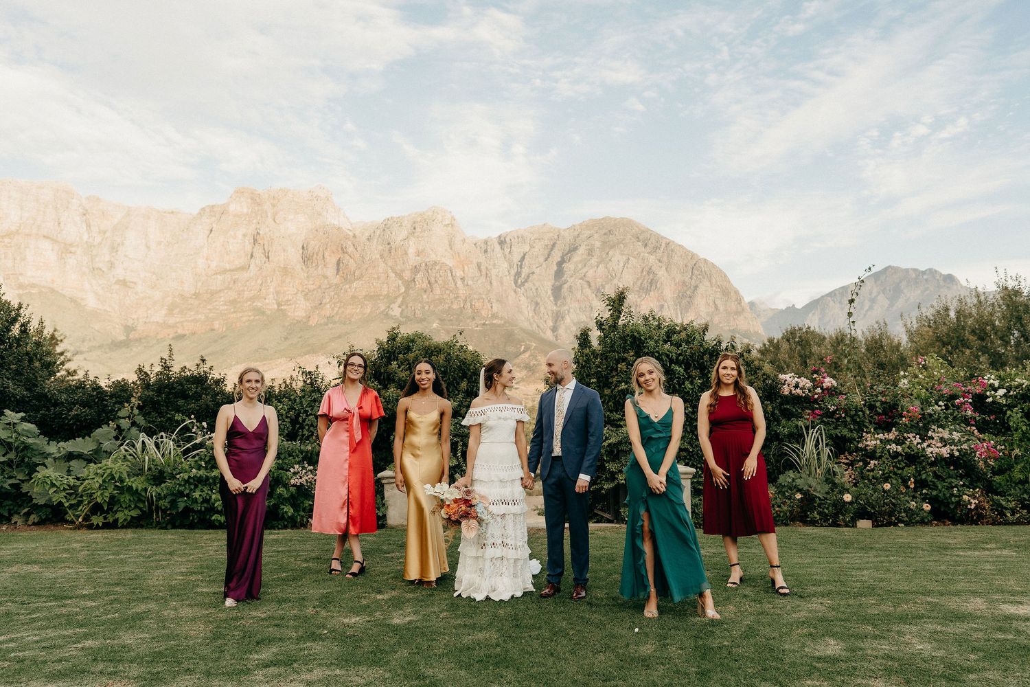 Wedding party stands on a lush lawn against dramatic mountain backdrop at an outdoor venue in warm evening light.