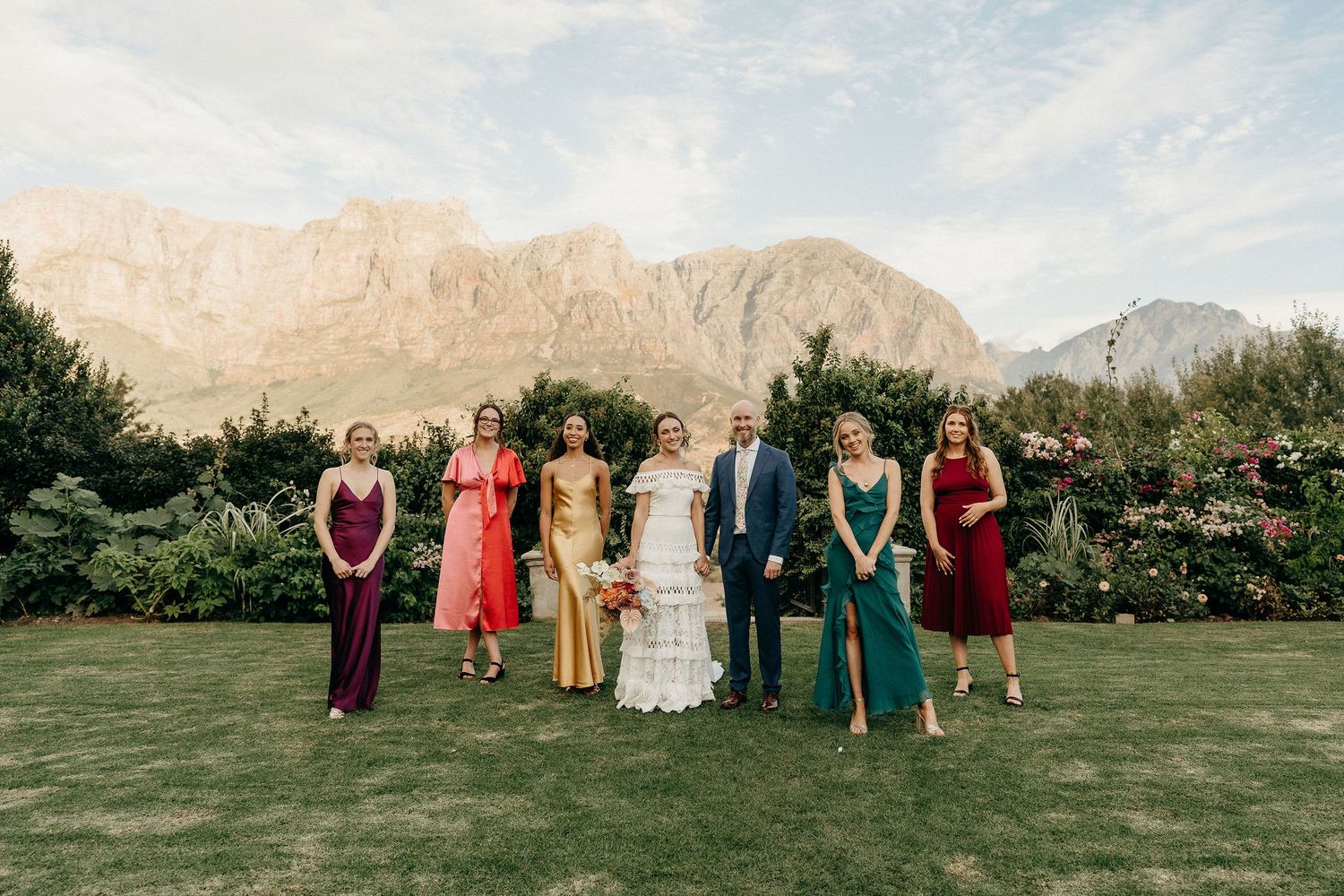 A wedding party stands on a manicured lawn with dramatic mountain peaks rising in the background.