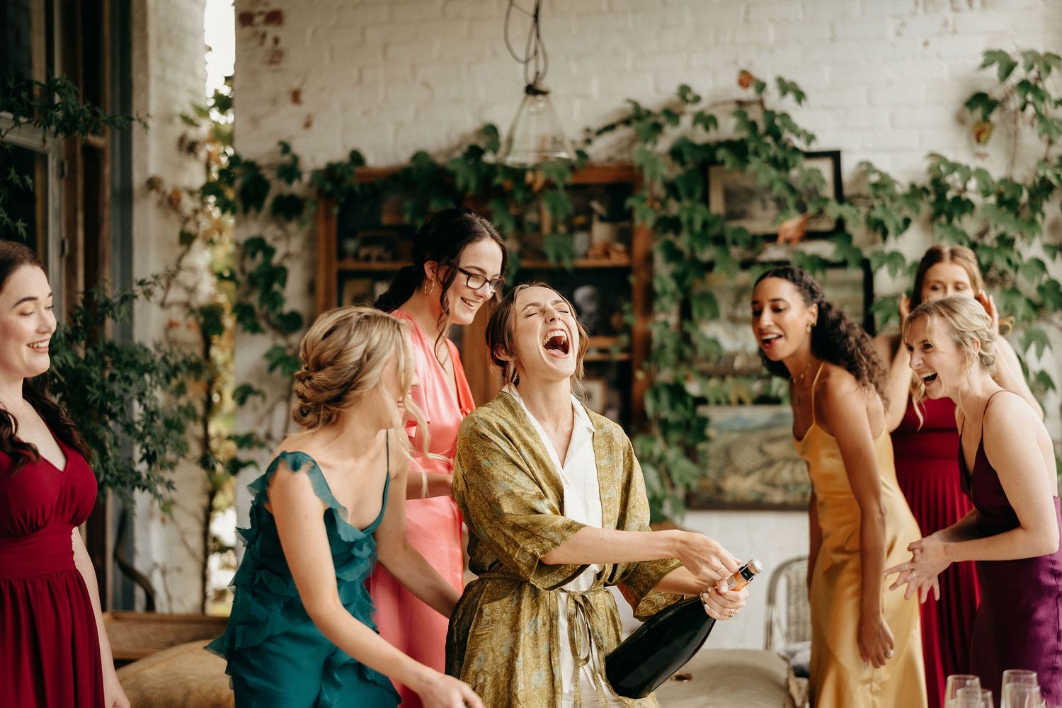 A group wearing colorful dresses and robes laughs joyfully together in a bright indoor space with climbing vines.