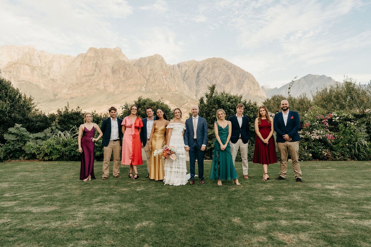 A group in formal attire stands on a lush lawn with dramatic mountain peaks rising behind them on a sunny day.