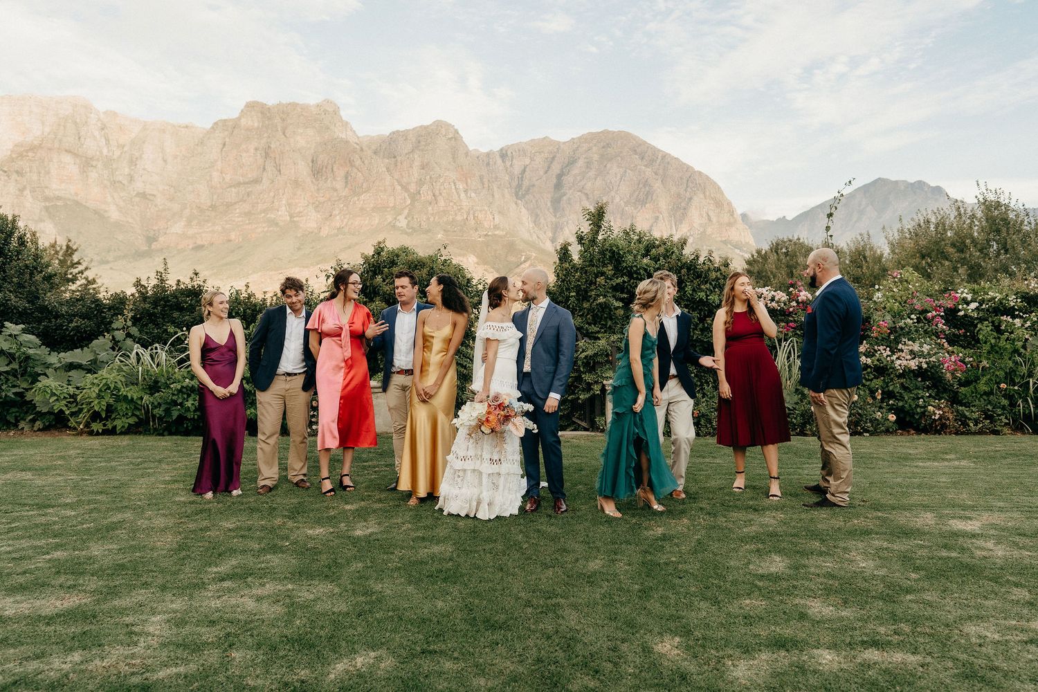 A group in formal attire stands on a lawn with majestic mountains creating a dramatic backdrop in the distance.